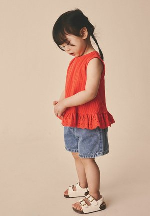 Malo dijete s pletenicama, odjeveno u crveni top bez rukava, traper kratke hlače i bijele sandale, gleda prema dolje s rukama sklopljenim.