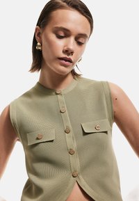 Olivgrön ärmlös topp med texturerad yta, med knappslå fram och två bröstfickor med matchande knappar.