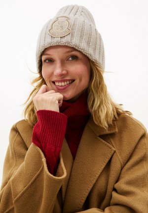 Lichtgrijze gebreide beanie met verticale ribbels en een ronde crèmekleurige logo patch. Model draagt een rode coltrui en een bruine jas.