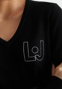 Pull noir à col en V avec un décolleté triangulaire et un logo en clous métalliques dans une police stylisée. Texture en tricot doux.