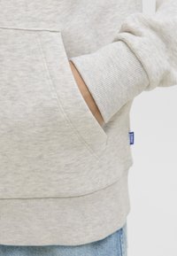 Roka v žepu svetlo sive hoodie s rebrastim manžetom, spodaj pa so delno vidne modre kavbojke.