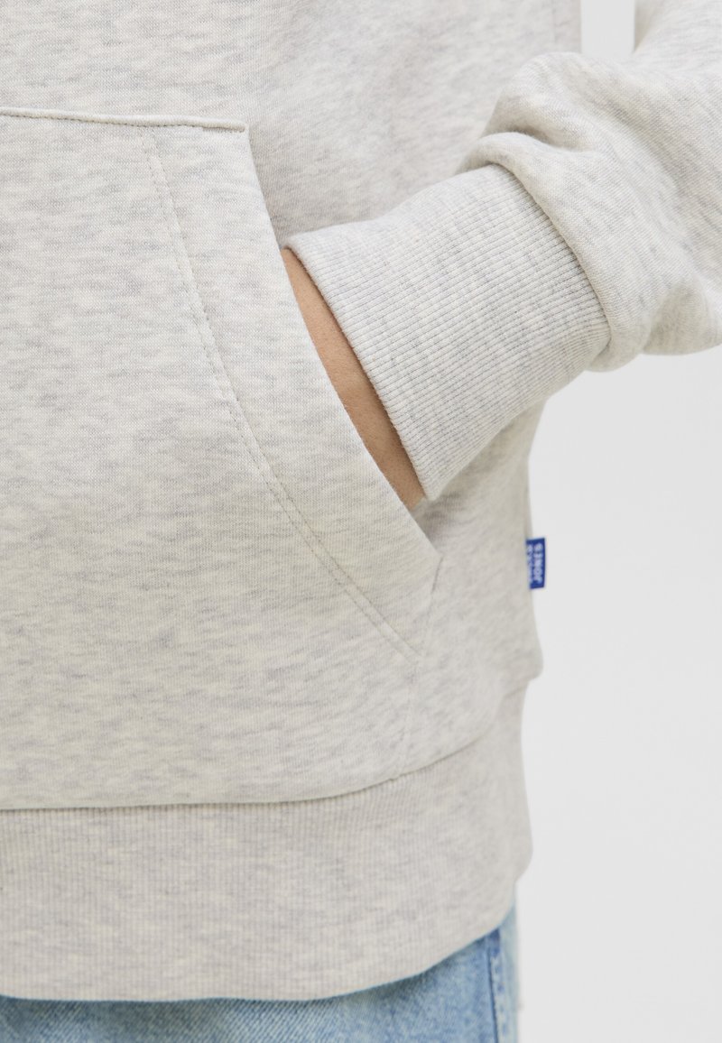 Roka v žepu svetlo sive hoodie s rebrastim manžetom, spodaj pa so delno vidne modre kavbojke.