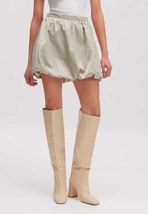 Model draagt een lichtgroene bubble-rok gecombineerd met beige kniehoge laarzen met hak en een wit mouwloos topje, staand op een effen achtergrond.