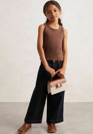 Mädchen mit langen Haaren in einem braunen ärmellosen Oberteil, weiten, dunklen Jeans, braunen Stiefeln, die eine beige und braune Handtasche mit goldenem Verschluss hält.