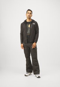 Felpa nera con zip e logo, t-shirt grafica nera e pantaloni cargo scuri. Indossa scarpe da ginnastica bianche. Design semplice, forma aderente.