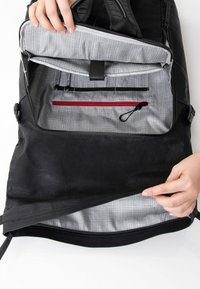 Zaino nero con un'esterno liscio e un interno a quadri grigi. Le caratteristiche includono zip, tasche e un compartimento principale accessibile.