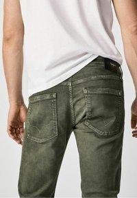 Green denim jeans with a slim fit, featuring stitched back pockets and a black label at the waistband, set against a plain background.