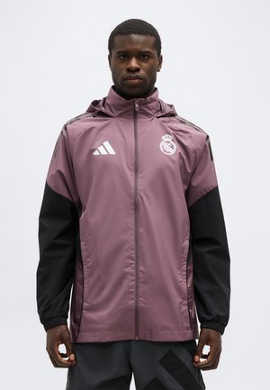 Hombre con chaqueta con cremallera de Real Madrid en púrpura y negro con el logo de Adidas, posando frente a un fondo claro y liso.