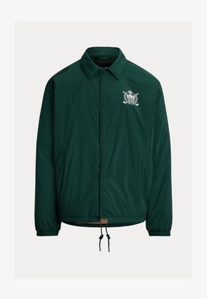 Tumma vihreä nappikiinnitteinen takki, jossa on kaulus, joustavat hihansuut, etutaskut, kiristyssuora helma ja valkoinen kirjailtu vaakuna rinnassa.