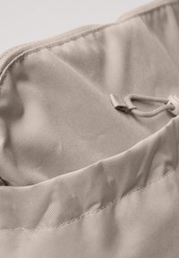 Nærbillede af beige stof med syninger og en lille løkke fastgjort til en snor eller snøre.