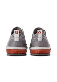 Zapatos de tela gris con un diseño texturizado, detalles en cuero gris y una suela de goma rojo-naranja. Presentan una etiqueta blanca con un logo.