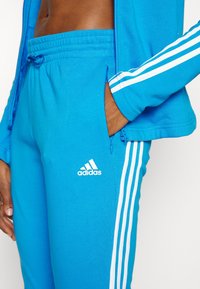 Personne portant un pantalon de survêtement Adidas bleu avec des bandes blanches et un logo, main dans une poche zippée, veste bleue partiellement visible avec des bandes.