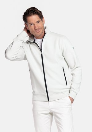Man met bruin haar, gekleed in een wit jack met rits en witte broek, poseert met één hand achter zijn hoofd en de andere in zijn broekzak.