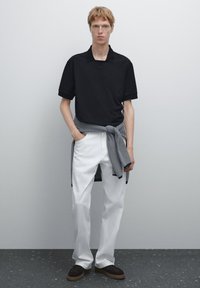 Černé polo tričko s krátkými rukávy spárované s bílými kalhotami. Proužkovaná černobílá tkanina uvázaná kolem pasu. Darkové boty dokončují tento outfit.