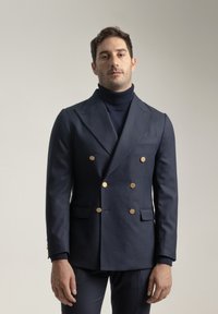 Blazer de doble botonadura en azul marino hecho de tela texturizada, con botones dorados, solapas estructuradas y bolsillos laterales. Lleva encima una blusa de cuello alto.