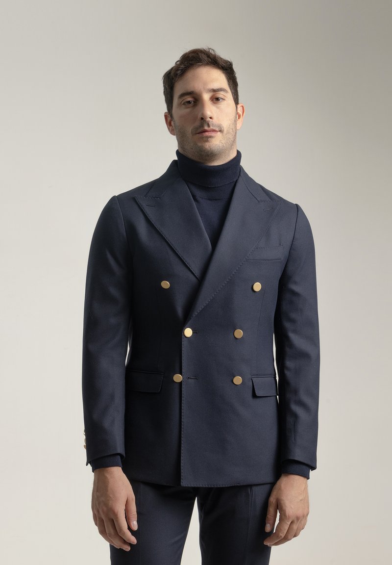 Blazer de doble botonadura en azul marino hecho de tela texturizada, con botones dorados, solapas estructuradas y bolsillos laterales. Lleva encima una blusa de cuello alto.