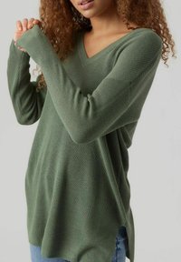 Modelka s kudrnatými vlasy má na sobě dlouhý, zelený svetr s výstřihem do V, který má ribovanou strukturu a uvolněné rukávy, na jednobarevném pozadí.