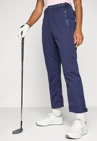 Osoba v námořnických golfových kalhotách a bílých teniskách svírá golfovou holi, což ukazuje na neformální, sportovní styl na neutrálním pozadí.