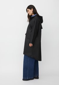 Donna con impermeabile nero con cappuccio, jeans larghi blu e scarpe nere, in piedi di lato contro uno sfondo bianco semplice.