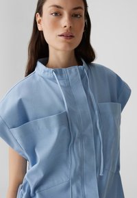 Lichtblauw, korte mouw top met een hoge kraag, twee voorzakken en een trekkoord. Gladde stof met minimale stikdetails.