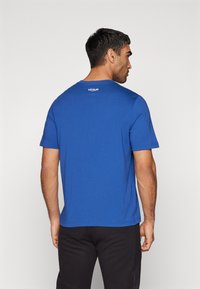Blå bomulds t-shirt med korte ærmer og rund halsudskæring. Har et lille hvidt logo på den øverste del af ryggen og et ensfarvet design.