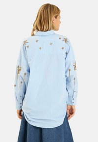 Lichtblauw langslavend shirt met gouden geborduurde bloemenpatronen, knopenmanchetten en een ronde zoom, ontworpen met een relaxed fit.