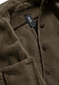 Cappotto in pelliccia sintetica marrone con una texture liscia, caratterizzato da un accento con catena, bottoni e un'etichetta. Il design include una grande tasca e un colletto.