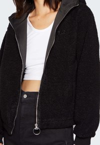 Czarna kurtka z polaru teddy z zamkiem błyskawicznym z przodu, szara podszewka kaptura i elastyczne mankiety. Posiada subtelne logo oraz zamek błyskawiczny z uchwytem w kształcie pierścienia.