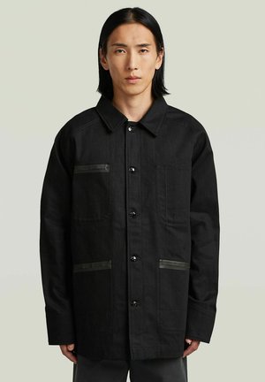 Person mit glatten schwarzen Haaren, die eine schwarz geknöpfte Jacke mit vier Vordertaschen vor einem schlichten hellen Hintergrund trägt.