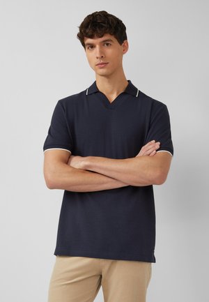 Mężczyzna ze skrzyżowanymi ramionami, ubrany w granatową, krótką koszulkę polo z białymi wykończeniami i beżowe spodnie, na jednolitym jasnym tle.