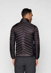 Melnā pūkainā jaka ar augstu apkakli, ar ševronu rakstu un šuvju detaļām. Aizmugures skats rāda gludu tekstūru un pieguļošu formu.