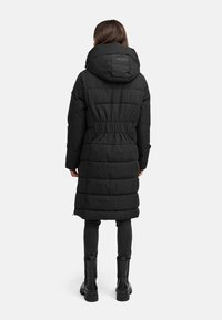 Langer schwarzer Daunenmantel mit tailliertem Schnitt, übergroßer Kapuze und gestepptem Muster. Kombiniert mit dunklen Leggings und klobigen schwarzen Stiefeln.