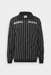 Svart vindjacka med vita vertikala ränder, med dragkedja fram, ståkrage och logotyp på bröstet i fet stil.