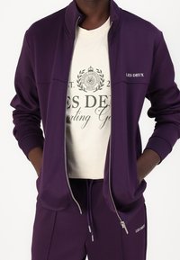 Chaqueta morada con cremallera, cuello alto, bolsillos laterales y puños acanalados, combinada con una camiseta gráfica crema con estampado verde y logotipo.