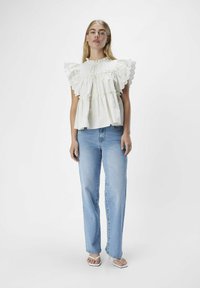 Top bianco arricciato con ampie maniche a scollatura ondulata, abbinato a jeans azzurri a gamba dritta e sandali bianchi. Design semplice, vestibilità casual.
