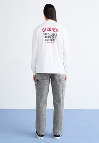 Cămașă albă cu mânecă lungă, cu text roșu și negru pe spate, combinată cu pantaloni cargo gri deschis, largi, cu buzunare.