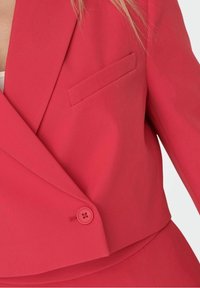 Blazer rojo hecho de tela suave, con cuello con muesca, cierre de un solo botón y un bolsillo en el pecho. Corte entallado con líneas limpias.