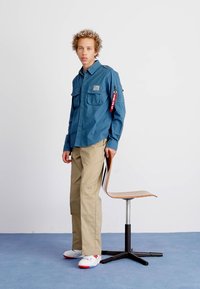 Modrá košeľa na gombíky s dvoma vreckami na hrudi, béžové cargo nohavice a biele topánky s červenými akcentami. Model stojí pri drevenej stoličke.