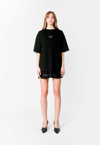 Camiseta negra de gran tamaño con mangas cortas, que presenta un pequeño parche con el logo. Combinada con pantalones cortos negros brillantes y tacones negros puntiagudos.