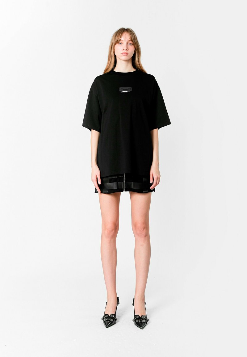 Camiseta negra de gran tamaño con mangas cortas, que presenta un pequeño parche con el logo. Combinada con pantalones cortos negros brillantes y tacones negros puntiagudos.