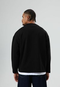 Zwarte sweatshirt met een losse pasvorm, voorzien van een ronde hals en verlaagde schouders. Draagt over een wit t-shirt, gecombineerd met donkere jeans.