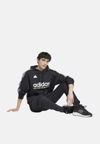 Felpa nera con logo "adidas SPORTSWEAR" bianco, caratterizzata da maniche con tre strisce. Abbinata a pantaloni neri e scarpe da ginnastica nere.