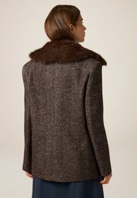 Brauner Mantel im Fischgrätenmuster mit einem plüschigen braunen Pelzkraagen. Maßgeschneiderte Passform und gerader Schnitt, mit langen Ärmeln und minimaler Detailgestaltung.