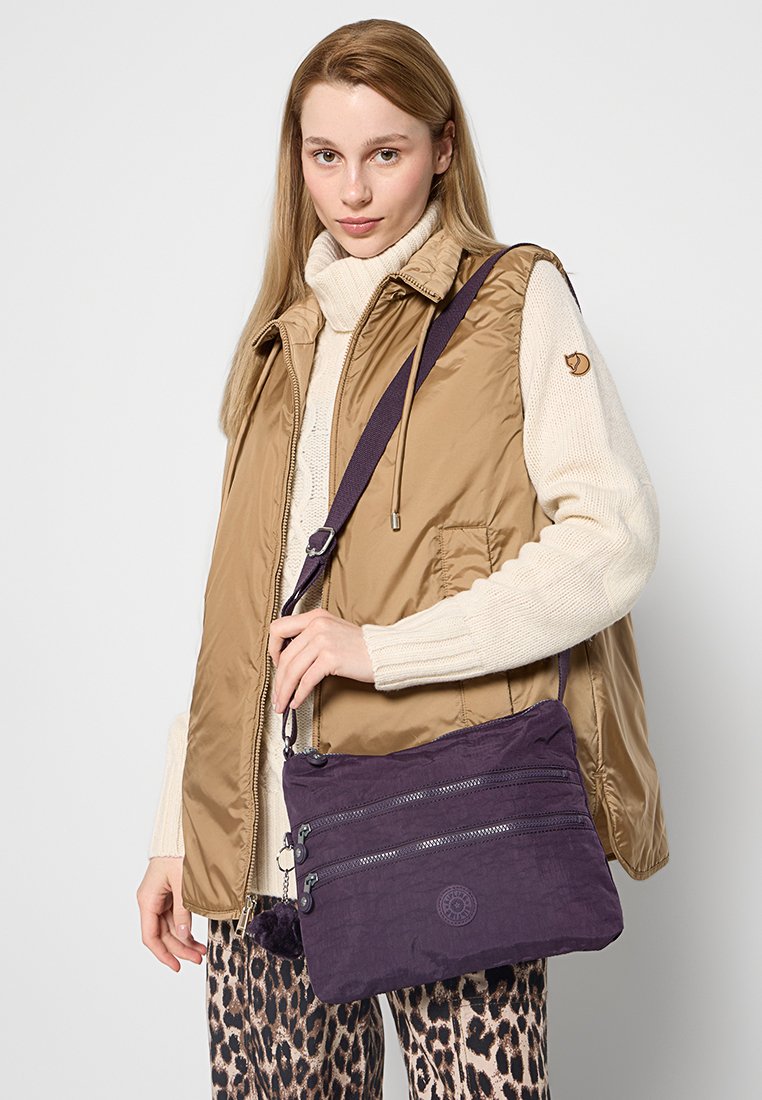 Borsa a tracolla di tessuto viola con tre scomparti con cerniera e un logo circolare, portata da un modello che indossa un gilet beige e pantaloni a fantasia.