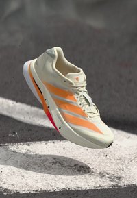 Hellbeiger Sportschuh mit orangefarbenen Akzenten, strukturiertem Mesh-Obermaterial, drei weißen Streifen und einer rosa Sohle. Leichtes Design zum Laufen.
