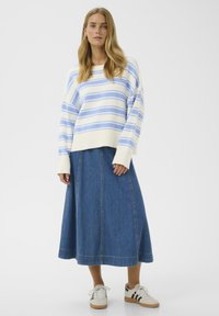 Femme portant un pull à rayures blanches et bleues, une longue jupe en denim bleu, et des baskets blanches avec des rayures noires, debout devant un fond blanc.