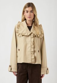 Jeune femme portant une veste à col à volants beige et un pantalon marron, debout devant un fond gris clair uni.