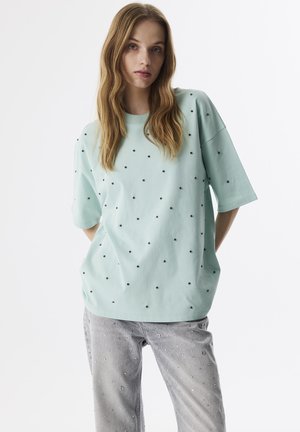 Sweat-shirt vert menthe à manches courtes, avec des pois noirs. Le tissu semble doux, et le design est décontracté et surdimensionné.