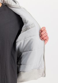 Světlé šedé podšívka zatepleného kabátu s měkkou strukturou, se stříbrným zipem a patentním knoflíkem. Viditelné šedé žebrované lemy a manžety.