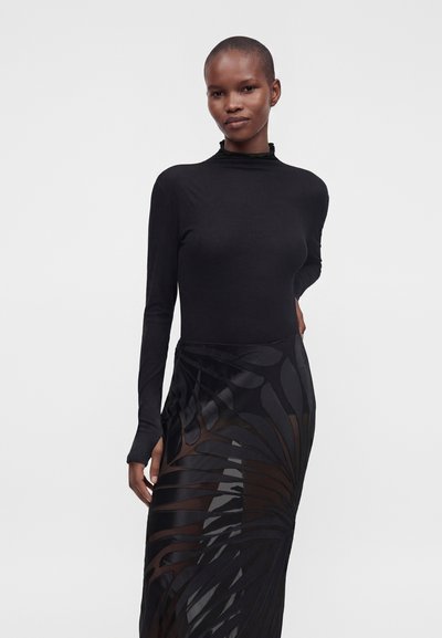 Personne portant un haut noir moulant à manches longues et une jupe noire transparente avec de larges motifs en forme de feuilles, debout devant un fond uni.